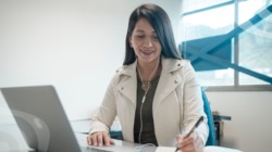 Una mujer sonriendo mientras trabaja con un computador y toma apuntes con un esfero