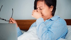 Una mujer, sentada en su cama y usando pijama, se ha retirado sus gafas y demuestra agotamiento visual. Sobre la cobija que cubre sus piernas, se encuentra un computador portátil abierto.