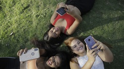 tres mujeres jóvenes acostadas en el césped alzan sus manos y juegan con sus dispositivos móviles